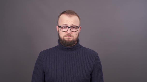 Portrait of a young bearded man in glasses, who asking for forgiveness. Feeling confused. Isolated on grey background. — Stock Video