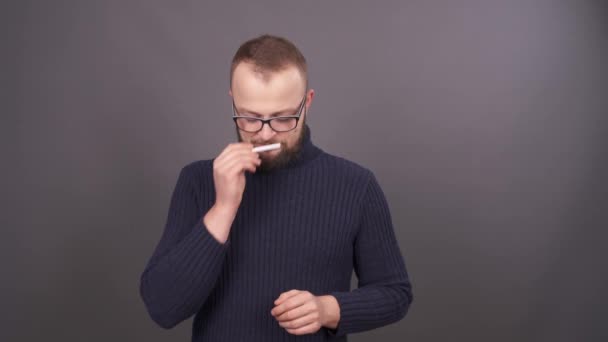 Porträtt av en ung skäggig man i glasögon, tar en cigarett från pocket, sniffa det, få glädje. Börjar hosta och döljer det tillbaka, då beslutar att röka. Isolerad på grå bakgrund. — Stockvideo