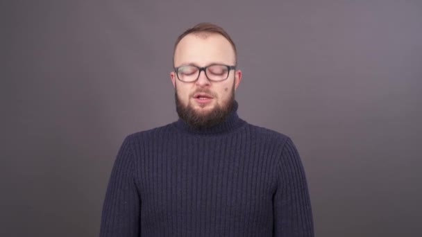 Närbild porträtt av en skäggig ung snotty kaukasiska man i glasögon, nysningar och blåser näsan. Isolerad på grå bakgrund. — Stockvideo