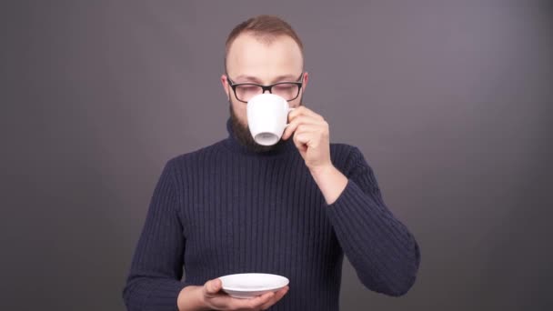 Retrato de cerca de un joven caucásico barbudo en vasos bebiendo café y sonriendo en cámara. Aislado sobre fondo gris . — Vídeos de Stock