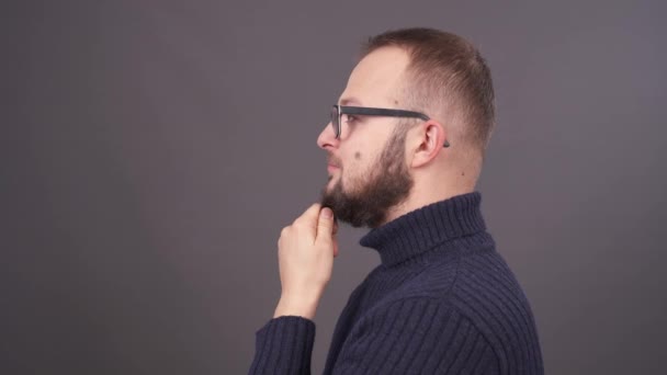 Retrato de cerca de un joven caucásico pensativo con gafas acariciando su barba. El hombre se quita las gafas y se las lleva a la boca. Aislado sobre fondo gris . — Vídeo de stock