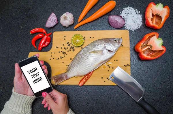 Young woman consulting recipe for fish on smartphone, fish and ingredients on the table. Preparing a gastronomic dish.