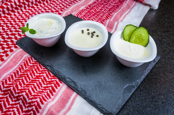 Delicious Lebanese (Arabic) food, thum, labneh and tzatziki sauces on black slate stone background and traditional Lebanese Keffiyeh turban.