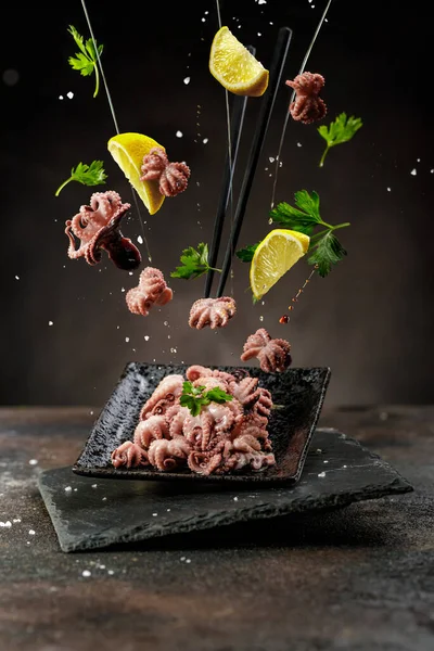 Levitated or flying food concept. Traditional Japanese food. Mini boiled ocopus, lemons, salt and parsley greens with chopsticks flying abovethe black grounge table.