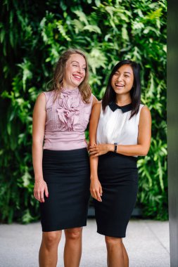 İki iş (bir Çinli Asyalı kadın ve beyaz bir kadın) farklı bir takım portresi yan yana bitkiler yeşil bir duvara karşı ayakta. Rahat bir şekilde gülümsüyor musun.