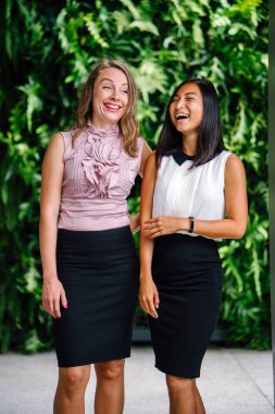 İki iş (bir Çinli Asyalı kadın ve beyaz bir kadın) farklı bir takım portresi yan yana bitkiler yeşil bir duvara karşı ayakta. Rahat bir şekilde gülümsüyor musun.