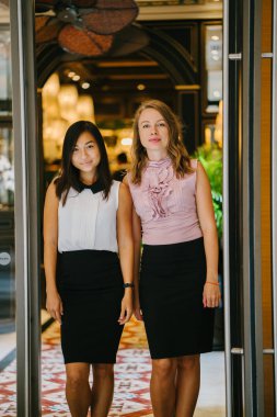 İki iş (bir Çinli Asyalı kadın ve beyaz bir kadın) farklı bir takım portresi yan yana bitkiler yeşil bir duvara karşı ayakta. Gülümseyen ve rahat bir şekilde gülerek.