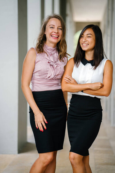 Portrait of a Chinese Asian woman and Caucasian white woman next to one another. They are both smiling  and are dressed professionally.