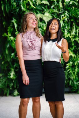 İki iş (bir Çinli Asyalı kadın ve beyaz bir kadın) farklı bir takım portresi yan yana bitkiler yeşil bir duvara karşı ayakta. Rahat bir şekilde gülümsüyor musun.