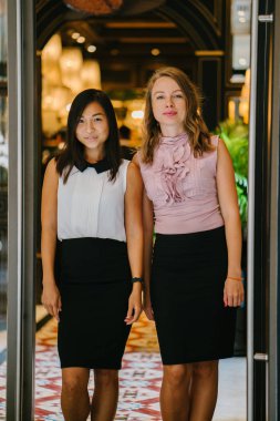 İki iş (bir Çinli Asyalı kadın ve beyaz bir kadın) farklı bir takım portresi yan yana bitkiler yeşil bir duvara karşı ayakta. Gülümseyen ve rahat bir şekilde gülerek.