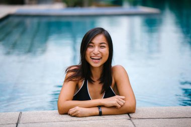 Genç bir Çinli Asyalı kız onun mayo gün boyunca bir havuz kenarına eğilerek portresi. O gülümseyen ve kamera için gülümsüyor gibi samimi ve rahat bir şekilde gülüyor.