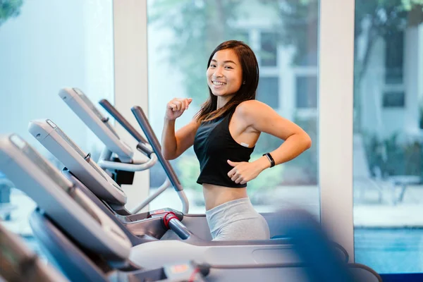 Chinese Asian woman is running on a treadmill in the gym. She is young, attractive, tanned and fit and is smiling as she enjoys her run.