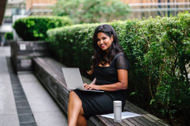 Portrait of a young Indian Asian woman sitting on a bench in the park during the day and working on her laptop with several documents. She is youthful but confident and mature and is focused on work. clipart