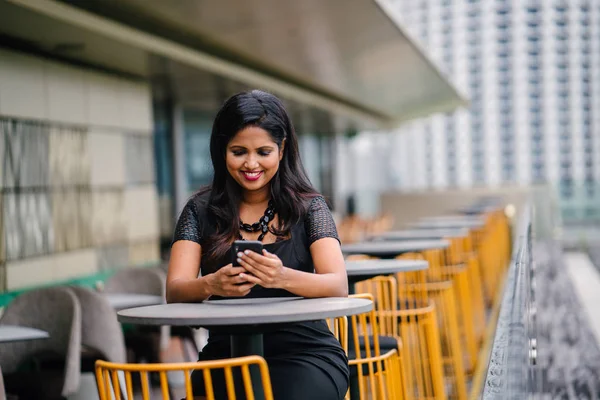 Portrait of a successful and confident Indian Asian business woman talking on her mobile smartphone in the city. She is sitting outdoors with Singapore city in the background and smiling as she talks.