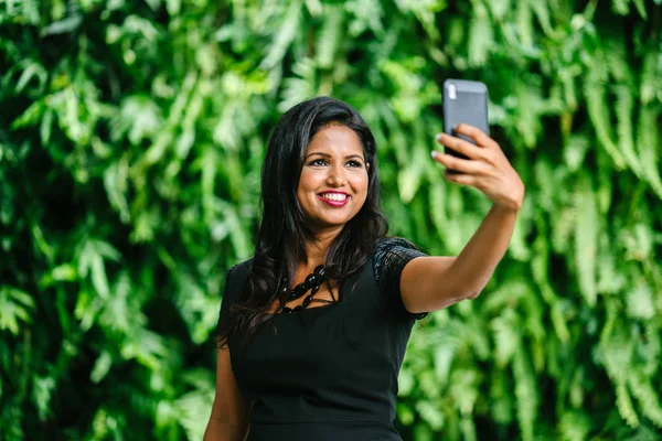 Portrait of a youthful but confident and mature Indian woman taking a selfie of herself with her cell phone against a green wall of plants in the day. She is smiling radiantly for her photo