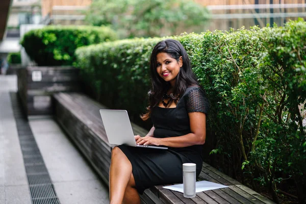 Portrait of a young Indian Asian woman sitting on a bench in the park during the day and working on her laptop with several documents. She is youthful but confident and mature and is focused on work.