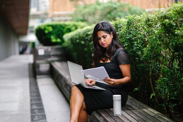 Portrait of a young Indian Asian woman sitting on a bench in the park during the day and working on her laptop with several documents. She is youthful but confident and mature and is focused on work.