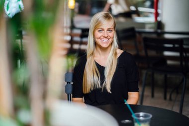  sarışın kadın bir kafede oturur ve bir içki ile gün boyunca rahatlatır genç ve güzel beyaz beyaz. Asya tropik bir şehirde değil. Gülümsüyor 