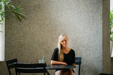  sarışın kadın bir kafede oturur ve bir içki ile gün boyunca rahatlatır genç ve güzel beyaz beyaz. Asya tropik bir şehirde değil. Gülümsüyor 