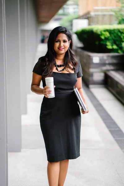 confident Indian Asian woman is walking down a corridor while she holds her laptop, documents and a flask of coffee. She is smiling as she strides down the shaded path in the day.