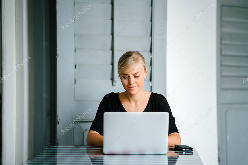 Portrait of a young and attractive blond woman from startup working at a desk during the day on her laptop. She is focused on her work and is typing on her notebook and has her hair tied up.