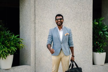 Günde bir duvara yaslanmış cool Hint Asya adam portresi. O sıradan bir takım elbise ve güneş gözlüğü. O güneşin altında uzanmanız ve birini bekliyor gibi bir spor çantası elinde.