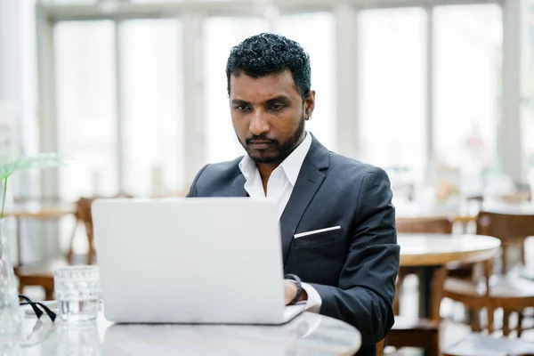 handsome and roguish Indian Asian man in a 3 piece suit is working on his laptop. He is seated at a table during the day and looks calm