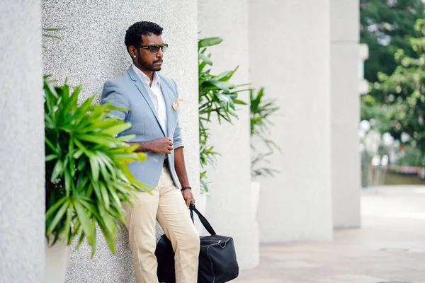 Günde bir duvara yaslanmış cool Hint Asya adam portresi. O sıradan bir takım elbise ve güneş gözlüğü. O güneşin altında uzanmanız ve birini bekliyor gibi bir spor çantası elinde.