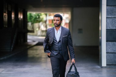 Portrait of a handsome Indian Asian man in a 3-piece suit and holding a bag walking down the street. He is silhouetted against the city and is walking confidently. clipart