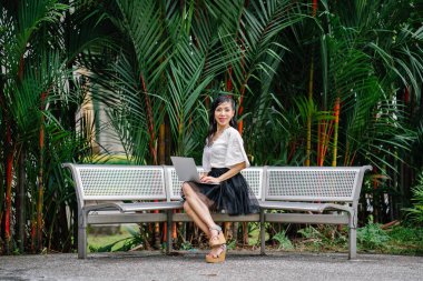 Asyalı bir kadın ağaçları içinde belgili tanımlık geçmiş ile metal bir bankta oturmuş ve dizüstü bilgisayarında çalışıyor. O orta yaşlı, ince ve ofis kıyafet giymiş.