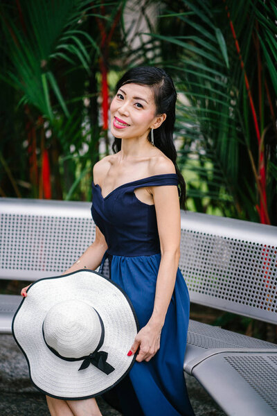 Portrait of Asian Chinese woman in an elegant blue dress posing with a white broad-brimmed hat during the day in a park in Asia.