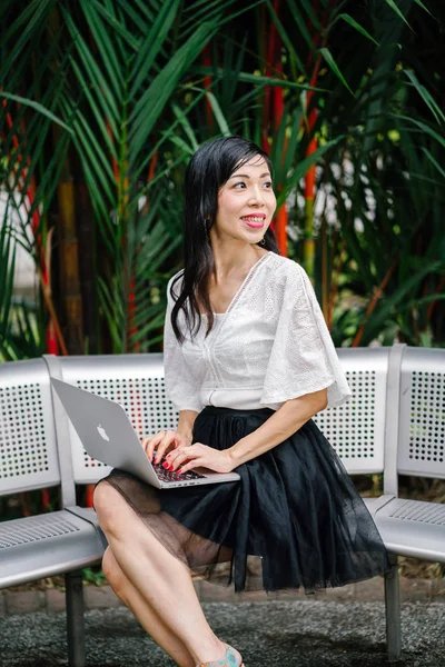 An Asian woman is sitting on a metal bench with trees in the background and is working on her laptop computer. She is middle aged, thin and is dressed in office attire.