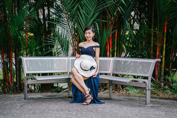 Portrait of Asian Chinese woman in an elegant blue dress posing with a white broad-brimmed hat during the day in a park in Asia.