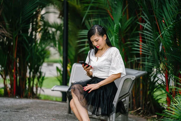 An Asian woman is sitting on a metal bench with trees in the background with smartphone. She is middle aged, thin and is dressed in office attire.