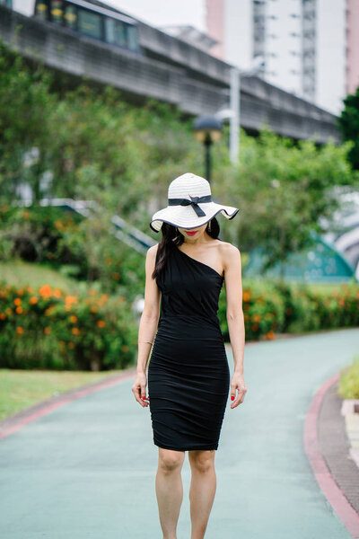 Portrait of Asian Chinese woman in an elegant  black dress posing with a white broad-brimmed hat during the day in a park in Asia.
