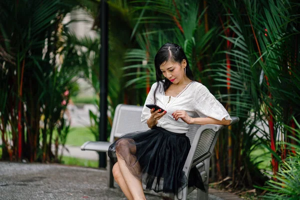 An Asian woman is sitting on a metal bench with trees in the background with smartphone. She is middle aged, thin and is dressed in office attire.