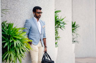 Günde bir duvara yaslanmış cool Hint Asya adam portresi. O sıradan bir takım elbise ve güneş gözlüğü. O güneşin altında uzanmanız ve birini bekliyor gibi bir spor çantası elinde.