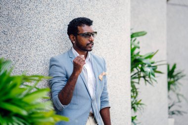 Günde bir duvara yaslanmış cool Hint Asya adam portresi. O sıradan bir takım elbise ve güneş gözlüğü. O güneşin altında uzanmanız ve birini bekliyor gibi bir spor çantası elinde.