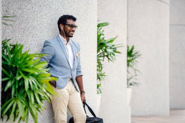 Günde bir duvara yaslanmış cool Hint Asya adam portresi. O sıradan bir takım elbise ve güneş gözlüğü. O güneşin altında uzanmanız ve birini bekliyor gibi bir spor çantası elinde.