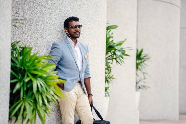 Günde bir duvara yaslanmış cool Hint Asya adam portresi. O sıradan bir takım elbise ve güneş gözlüğü. O güneşin altında uzanmanız ve birini bekliyor gibi bir spor çantası elinde.