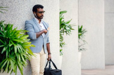 Günde bir duvara yaslanmış cool Hint Asya adam portresi. O sıradan bir takım elbise ve güneş gözlüğü. O güneşin altında uzanmanız ve birini bekliyor gibi bir spor çantası elinde.