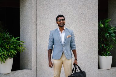 Günde bir duvara yaslanmış cool Hint Asya adam portresi. O sıradan bir takım elbise ve güneş gözlüğü. O güneşin altında uzanmanız ve birini bekliyor gibi bir spor çantası elinde.