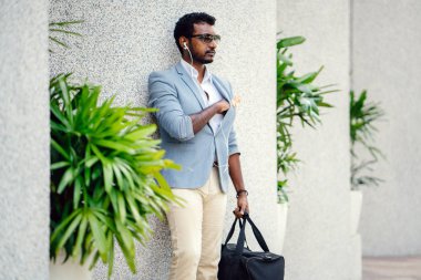 Günde bir duvara yaslanmış cool Hint Asya adam portresi. O sıradan bir takım elbise ve güneş gözlüğü. O güneşin altında uzanmanız ve birini bekliyor gibi bir spor çantası elinde.