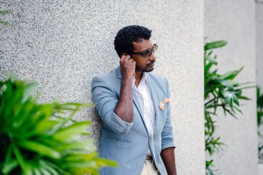 Günde bir duvara yaslanmış cool Hint Asya adam portresi. O sıradan bir takım elbise ve güneş gözlüğü. O güneşin altında uzanmanız ve birini bekliyor gibi bir spor çantası elinde.