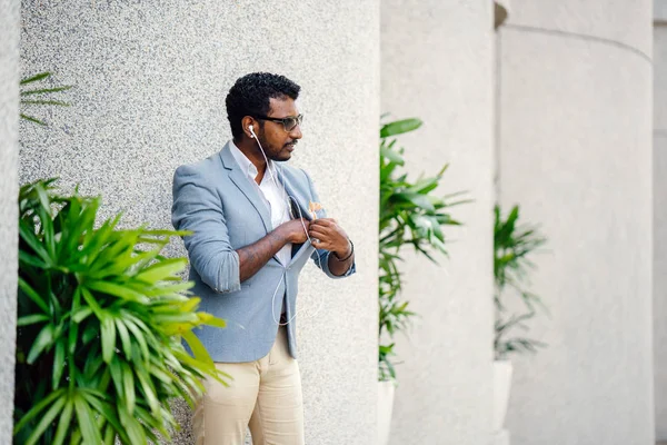 Günde bir duvara yaslanmış cool Hint Asya adam portresi. O sıradan bir takım elbise ve güneş gözlüğü. O güneşin altında uzanmanız ve birini bekliyor gibi bir spor çantası elinde.