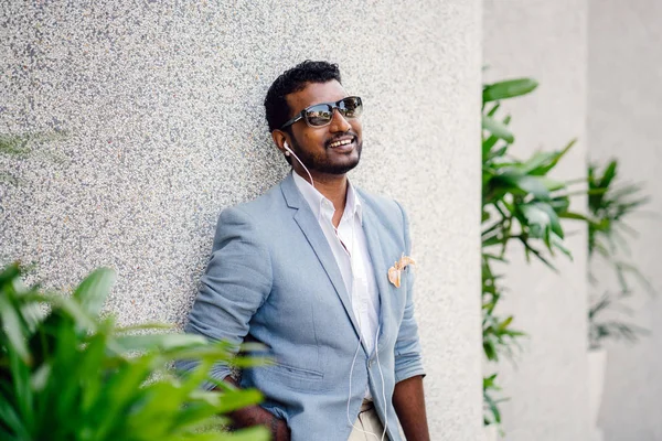 Günde bir duvara yaslanmış cool Hint Asya adam portresi. O sıradan bir takım elbise ve güneş gözlüğü. O güneşin altında uzanmanız ve birini bekliyor gibi bir spor çantası elinde.