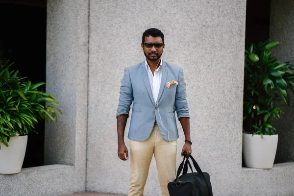 Portrait of a cool Indian Asian man leaning against a wall in the day. He is in a casual suit and sunglasses. He is holding a gym bag as he is lounging in the sun and waiting for someone.