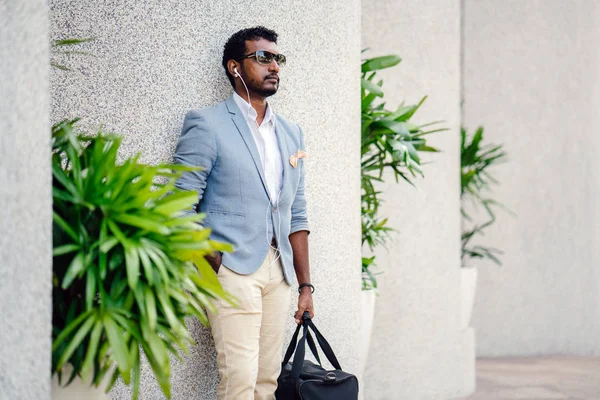 Günde bir duvara yaslanmış cool Hint Asya adam portresi. O sıradan bir takım elbise ve güneş gözlüğü. O güneşin altında uzanmanız ve birini bekliyor gibi bir spor çantası elinde.