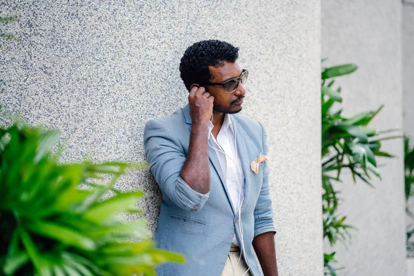 Günde bir duvara yaslanmış cool Hint Asya adam portresi. O sıradan bir takım elbise ve güneş gözlüğü. O güneşin altında uzanmanız ve birini bekliyor gibi bir spor çantası elinde.