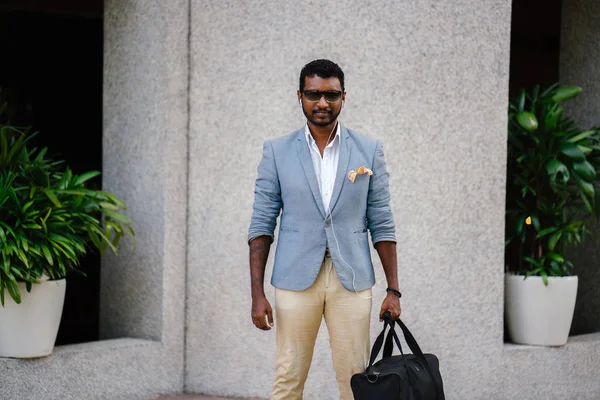 Portrait of a cool Indian Asian man leaning against a wall in the day. He is in a casual suit and sunglasses. He is holding a gym bag as he is lounging in the sun and waiting for someone.